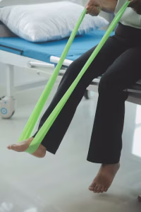 A cropped, close-up photo of a person seated on a medical examination table. They are holding a light green resistance band, which is looped around their bare feet.