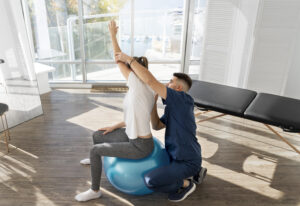 A physical therapist assists a person sitting on an exercise ball in the comfort of a bright home with large windows.