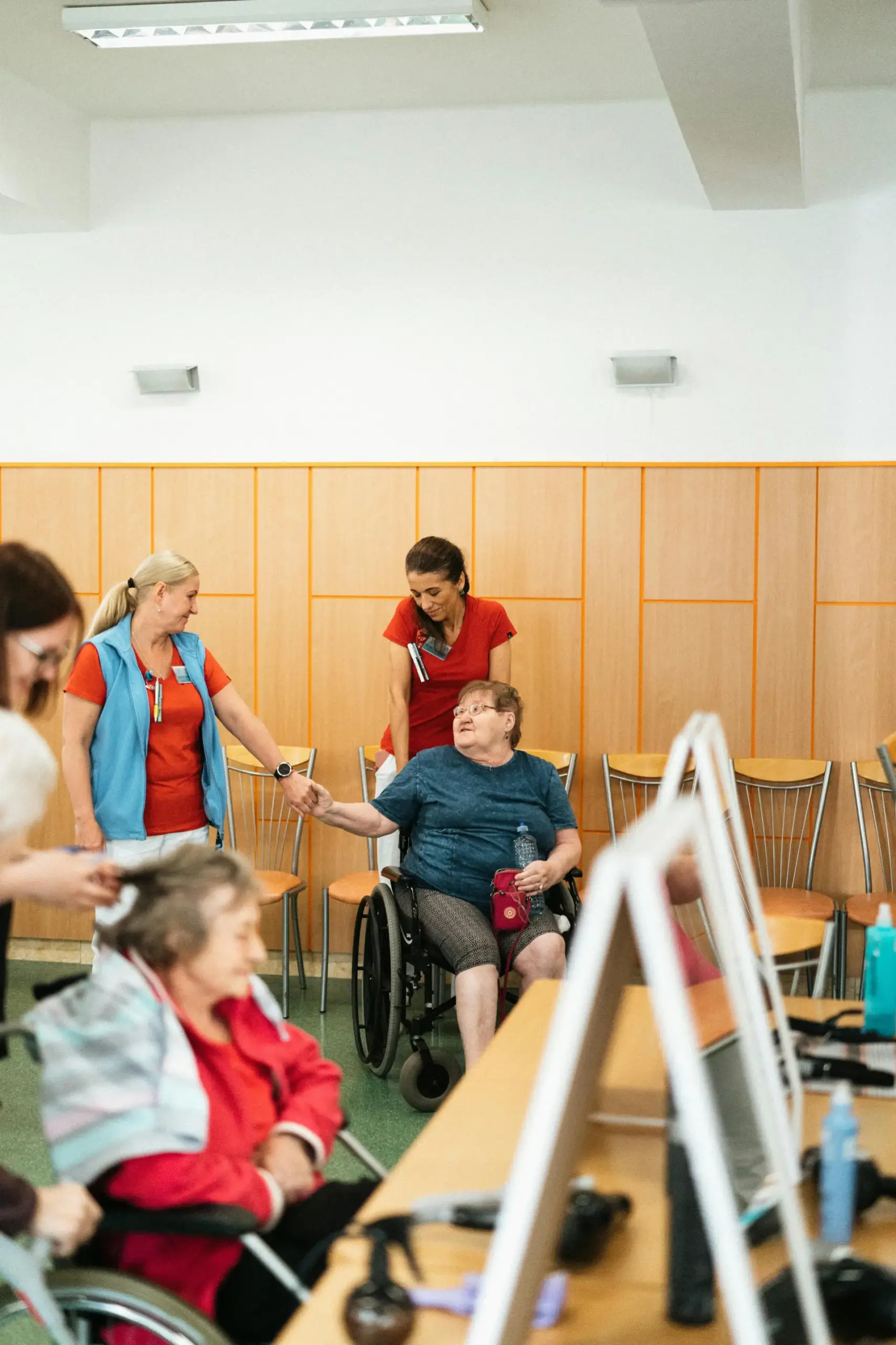 People gather in a room, some seated and others standing, including individuals with wheelchairs, as a mobile physiotherapy session brings the care directly to them.